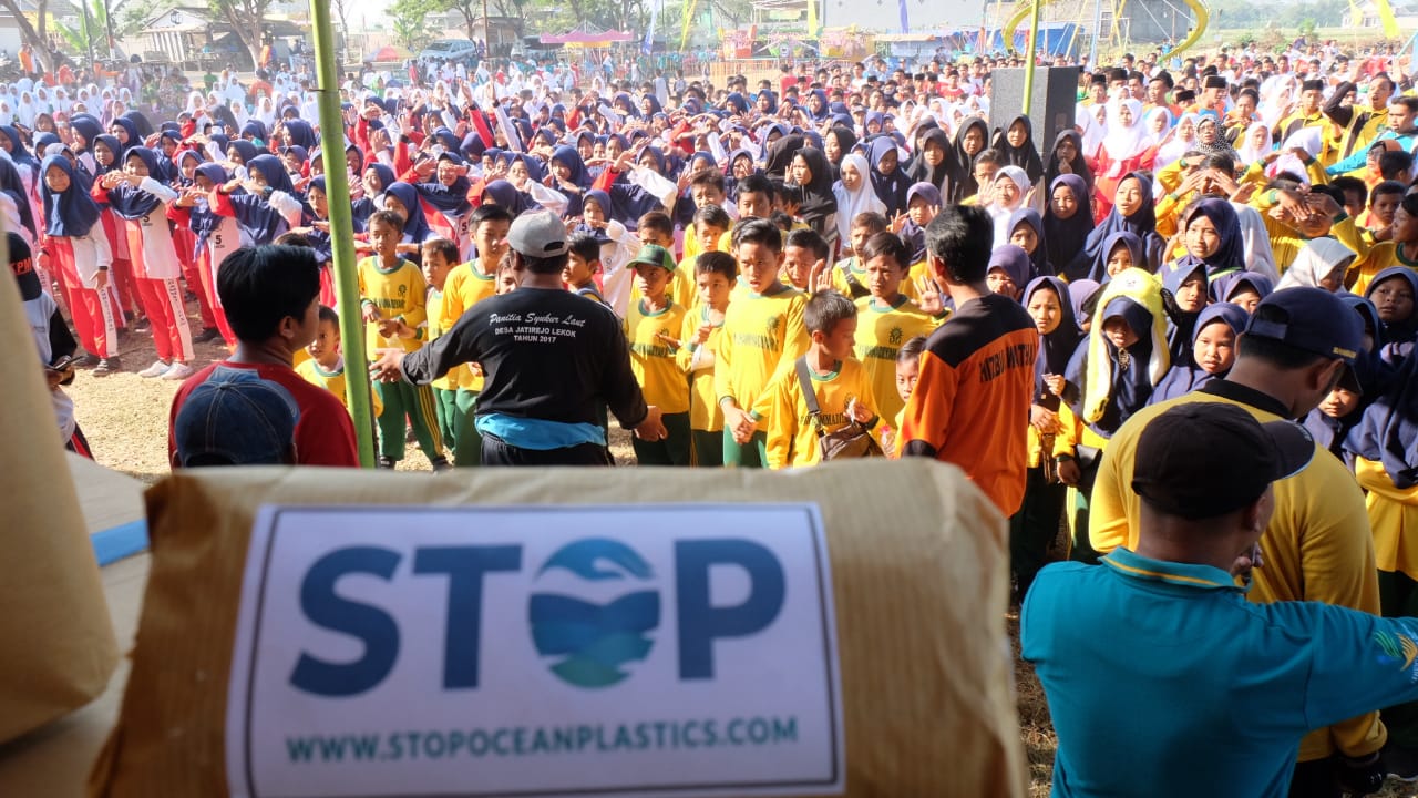Large community event in coastal Indonesia with hundreds of attendees, schoolchildren in uniform and a banner reading 